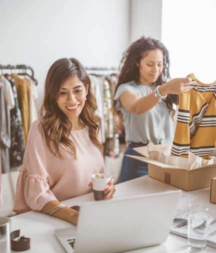 women working at clothing store. one on laptop, one looking at a shirt