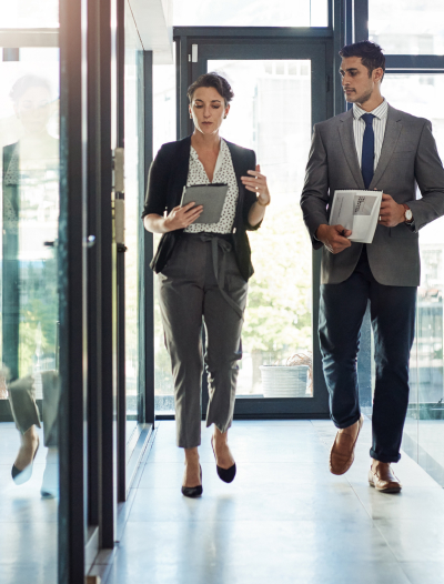 Two business people walking down an office hallway, discussing while looking at a tablet.